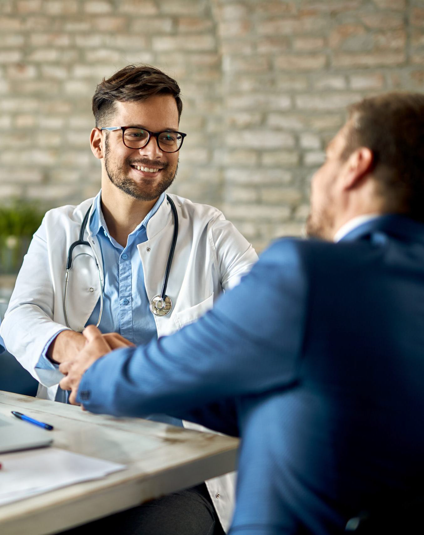 A smiling doctor wearing glasses and a white coat shakes hands with a man in a suit across a desk, illustrating positive Converged Provider Enablement in a friendly, professional setting.