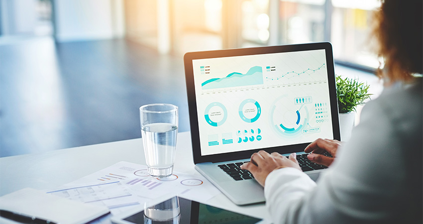 A person works on a laptop displaying graphs and charts, likely analyzing data related to artificial intelligence in healthcare. A glass of water, documents, a smartphone, and a small plant are on the desk in a bright office setting.