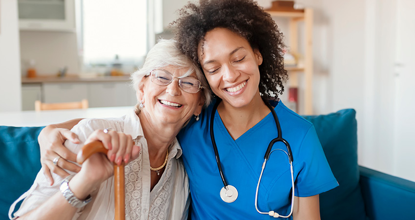 A smiling elderly woman with glasses and a cane sits beside a young healthcare worker in blue scrubs, both looking happy and relaxed on a sofa in a bright room, reflecting the compassionate care supported by SNF regulations.