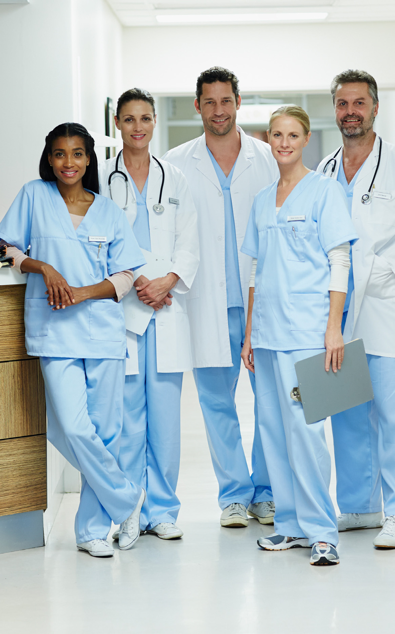 Five healthcare professionals in light blue scrubs and white lab coats stand together in a hospital hallway, smiling at the camera. Two hold clipboards, and two have stethoscopes around their necks.