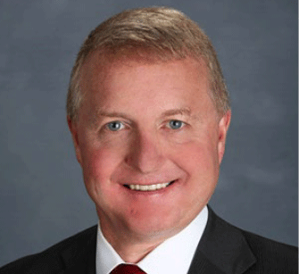 A middle-aged man with short, light brown hair is smiling. He is wearing a dark suit, white shirt, and red tie against a plain, gray background.