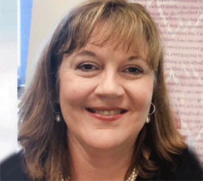 A woman with shoulder-length light brown hair and bangs is smiling. She is wearing pearl earrings and a pearl necklace, with a blurred background featuring some pink text.