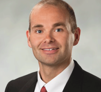 A man with short light brown hair, wearing a black suit, white shirt, and red tie, smiles at the camera against a plain, light-colored background.