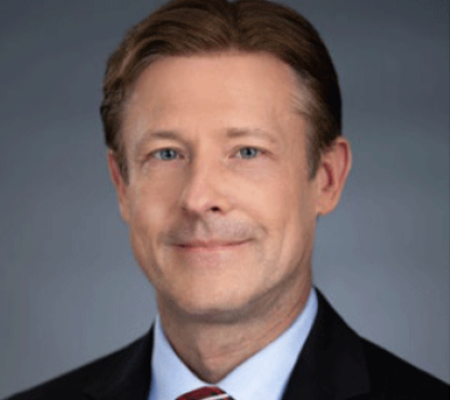 A man with light brown hair wearing a dark suit, white shirt, and striped tie, smiling slightly against a plain gray background.