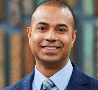 A man with short hair, wearing a blue suit jacket, light blue shirt, and a green striped tie, smiles at the camera. The background is blurred, showing vertical lines and muted colors.