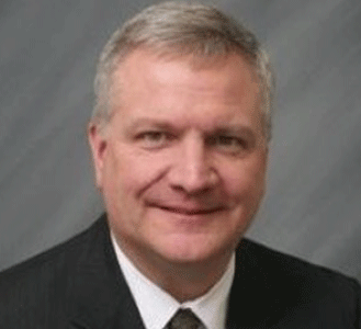 A middle-aged man with short gray hair, wearing a dark suit, white shirt, and tie, smiling in front of a plain gray background.
