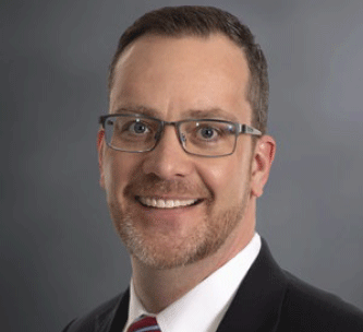 A man with short brown hair, glasses, and a trimmed beard, wearing a dark suit, white shirt, and striped tie, smiling at the camera against a plain gray background.