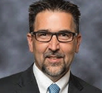 A man with short dark hair and a goatee is wearing glasses, a dark suit, a white shirt, and a light blue tie. He is smiling slightly and is posed in front of a gray, blurred background.