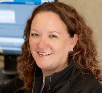 A woman with curly brown hair, wearing a black jacket and small hoop earrings, smiles at the camera. She is seated in front of a computer screen in an office setting.