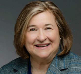 A middle-aged woman with shoulder-length light brown hair smiles warmly. She is wearing a blue plaid blazer and a dark top, with a neutral gray background behind her.