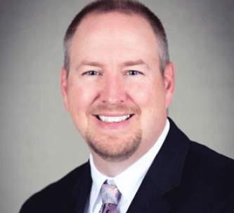 A man with short light hair and a goatee smiles at the camera. He is wearing a dark suit, white shirt, and patterned tie, posed against a plain, light-colored background.