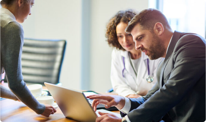 A man in a suit and a female doctor with a stethoscope are seated at a table, looking at a laptop screen, discussing Converged Provider Enablement, while another woman stands nearby, engaged in the conversation.