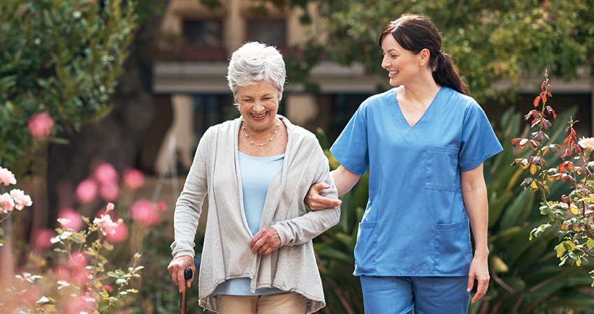 An elderly woman with gray hair uses a cane while walking outdoors, smiling, assisted by a younger woman in blue scrubs—demonstrating the caring support seen in SNF quality reporting. They are surrounded by greenery and flowers.