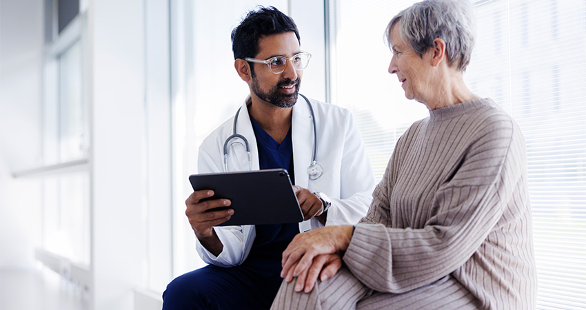 A doctor holding a tablet speaks with an older woman in a beige sweater about PCAST recommendations. They are sitting near large windows in a bright, modern medical office, sharing a friendly and supportive conversation.