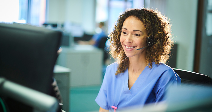 A smiling woman wearing medical scrubs and a headset sits at a computer in a brightly lit office, appearing to assist someone over the phone with the medical billing process.