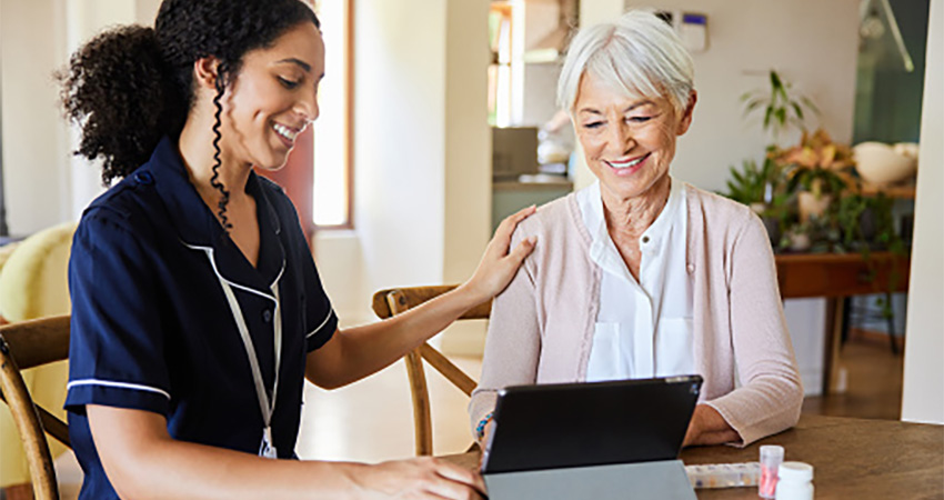 A young healthcare worker in uniform sits with an older woman at a table, smiling and looking at a tablet together as they explore autodetection in healthcare. The healthcare worker has a hand gently on the older woman's shoulder.