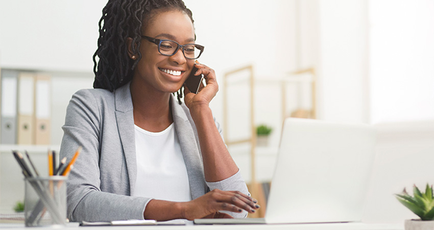 A smiling woman wearing glasses and a blazer sits at a desk, talking on the phone about changing patient insurance coverage while using a laptop in a bright, modern office setting.