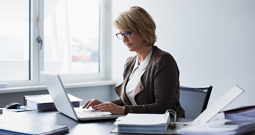 A woman with short blonde hair and glasses is sitting at a desk, typing on a laptop. She is dressed in business attire and surrounded by utilization management documents and binders in a bright office with a large window.