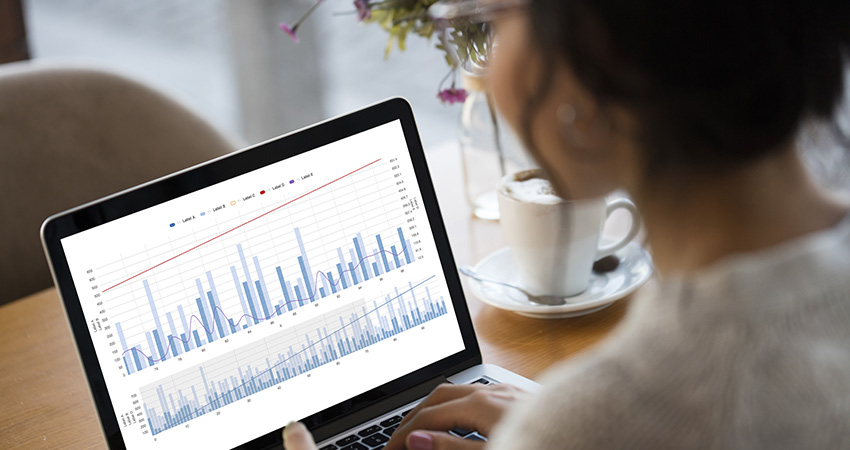A person sits at a table with a coffee cup, looking at a laptop displaying blue bar and red line graphs, analyzing data or statistics to refine their market access strategy.