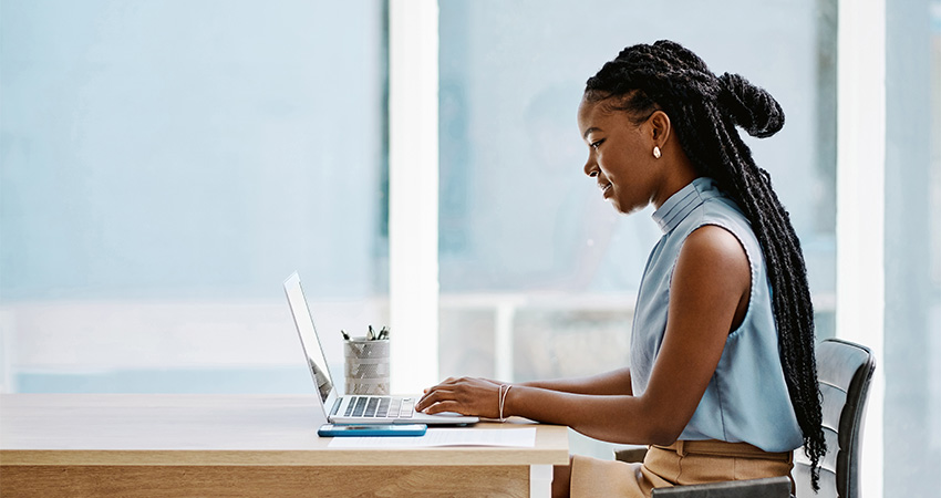 A woman with braided hair sits at a desk, smiling as she types on her laptop. Dressed in a light blue sleeveless top, she appears focused on data analytics work, with a pencil holder and notebook neatly arranged in front of her.