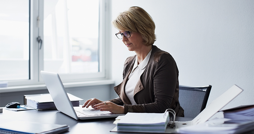 A woman with short blonde hair and glasses works on a laptop at a desk in a bright office, surrounded by documents and binders related to claims denials.