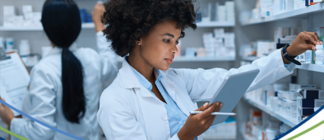 Two pharmacists in white lab coats work in a pharmacy; one checks shelves with medication while holding a tablet, and the other writes on a clipboard in front of stocked shelves.