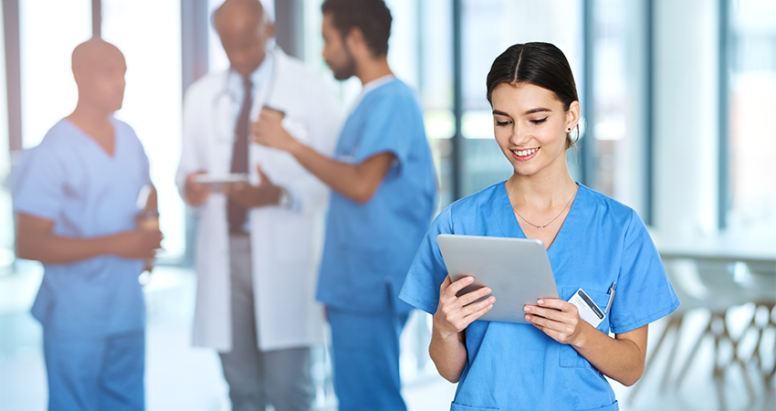 A smiling healthcare worker in blue scrubs uses the Inovalon Application Portal on a tablet in the foreground, while three medical professionals, including one in a white coat, have a discussion in the blurred background.