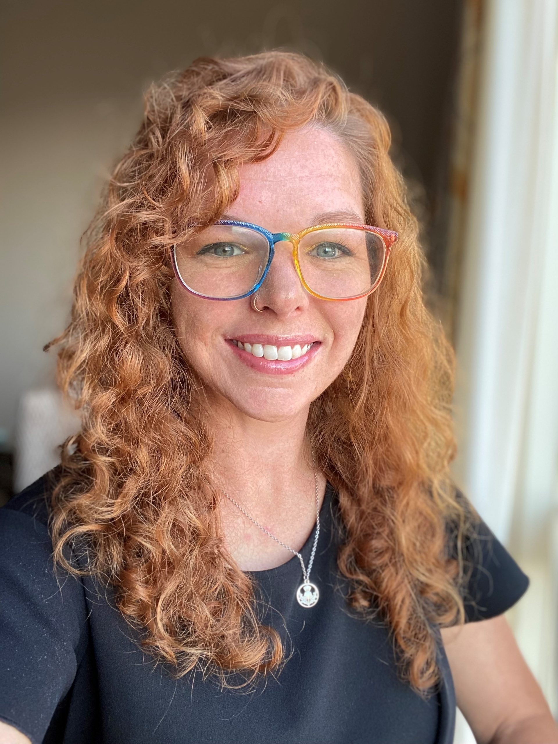 A woman with curly red hair and rainbow-rimmed glasses smiles at the camera. She is wearing a black top and a silver necklace, standing indoors near a window with sunlight illuminating her face.
