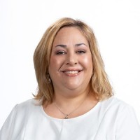 A woman with shoulder-length blonde hair wearing a white top smiles at the camera against a plain white background.