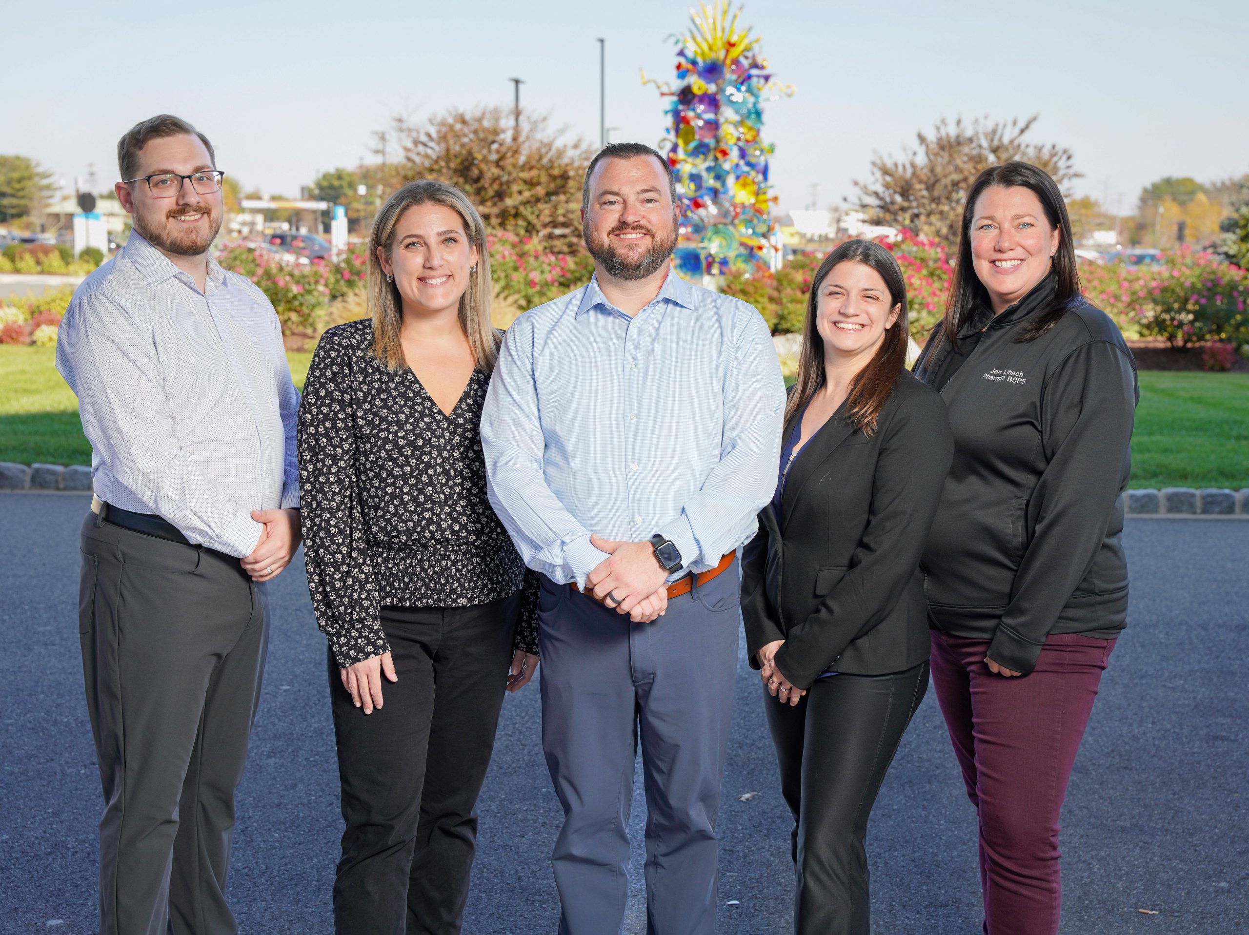 Five adults stand outdoors in a row, smiling at the camera. Behind them are green bushes, a colorful glass sculpture, and a clear sky. They are dressed in business casual attire.