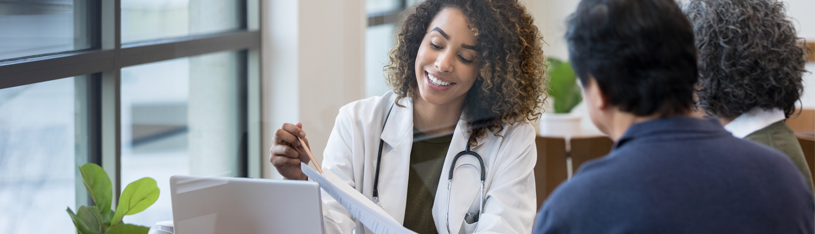 A smiling doctor with curly hair discusses liver cancer paperwork with two patients in an office. The doctor holds a file and wears a white coat and stethoscope. They sit at a desk near large windows and green plants.