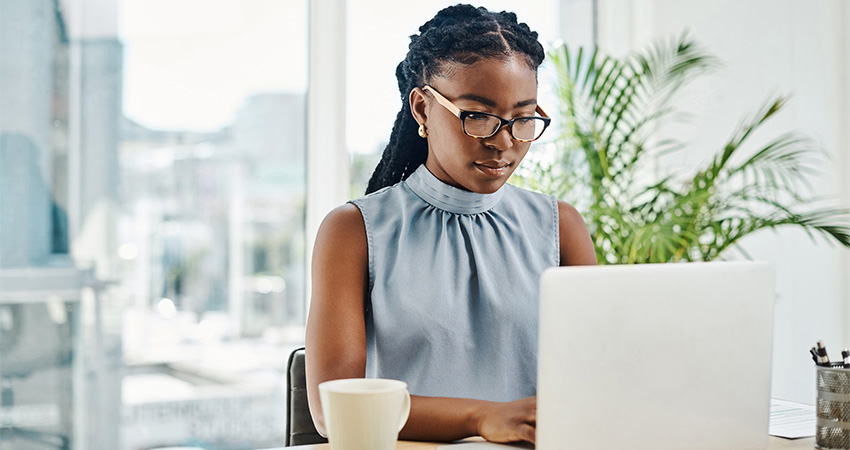 A woman wearing glasses and a sleeveless blouse works on a laptop at her desk, possibly reviewing HEDIS MY2026 Public comment. A coffee mug sits beside her, with a large green plant and a bright window in the background.