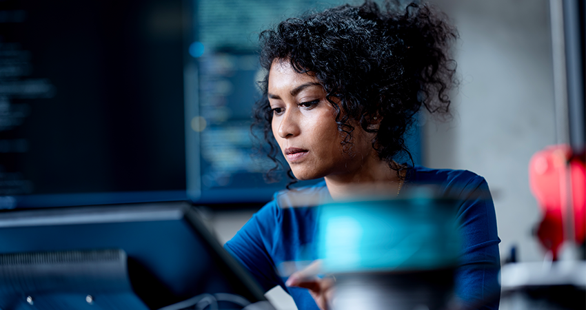 A woman with curly hair sits at a desk, focused on a computer screen. Blurred code on a monitor hints she is working on a technical task, possibly exploring the impact of AI in careers.