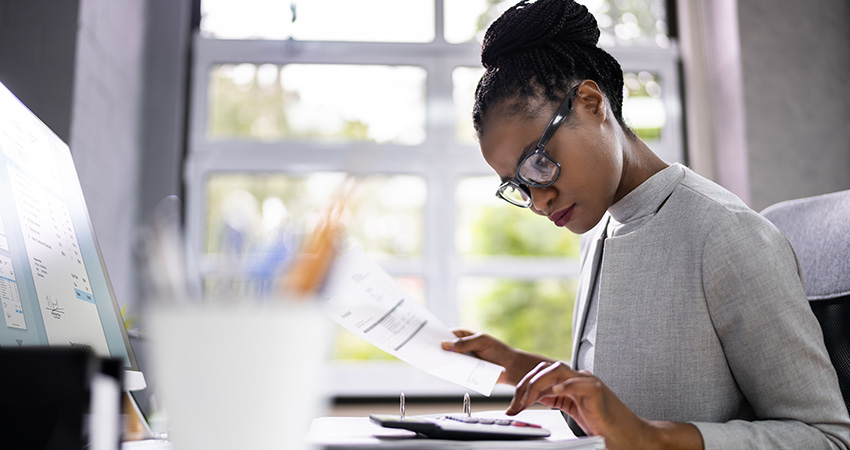 A woman wearing glasses sits at a desk, focused on medical billing for small practices as she reviews paperwork and uses a calculator. A computer monitor, office supplies, and a large window are visible in the background.