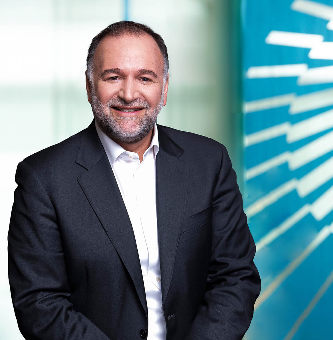 A man with short dark hair and a beard, wearing a dark suit and white shirt, smiles as he stands in front of a bright, modern blue and white geometric background, ready to improve healthcare outcomes.