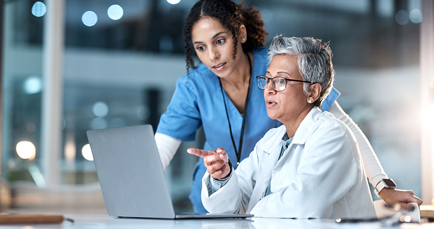 Two healthcare professionals, one in blue scrubs and one in a white lab coat, are reviewing big data analytics in healthcare on a laptop screen together in a modern medical office. The person in the lab coat is pointing at the screen.