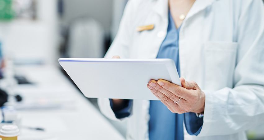 A medical professional in a white lab coat uses a digital tablet powered by Artificial Intelligence in Healthcare, standing in a clinical setting with medicine bottles slightly visible on the nearby counter.