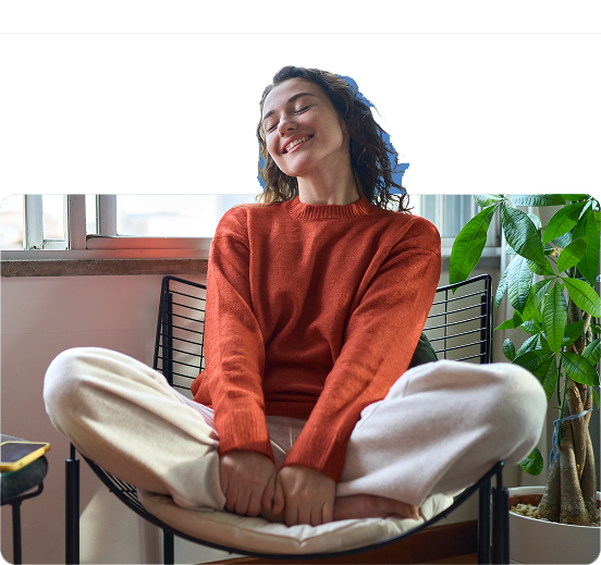 A smiling person in a red sweater and white pants sits cross-legged on a chair indoors, appearing peaceful—much like the seamless experience offered by the Inovalon ONE Platform. A large green plant and a window are in the background.