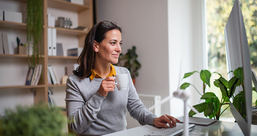 A woman sits at a desk in a bright home office, holding a white mug and smiling at her computer monitor, which displays MatrixCare. Bookshelves and green plants create a lively background.