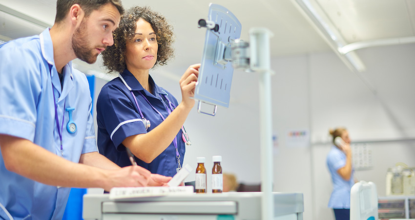 Two healthcare professionals in scrubs work at a computer station in a hospital. One types while the other writes on a clipboard, possibly exploring what is population health analytics. Medicine bottles and another person are visible in the background.