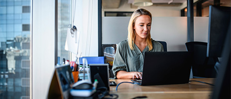 A woman sits at a desk in a modern office, working on a laptop. She is surrounded by office supplies and large windows with a view of city buildings outside.