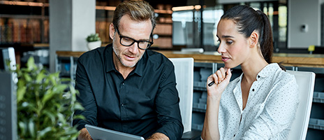 A man in glasses and a black shirt discusses something on a tablet with a woman in a white blouse, who is holding a pen. They are seated together in a modern, bright office space.