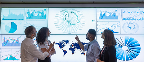 Four people stand in front of large screens displaying various charts, graphs, and a world map, discussing and analyzing the data visualizations together.