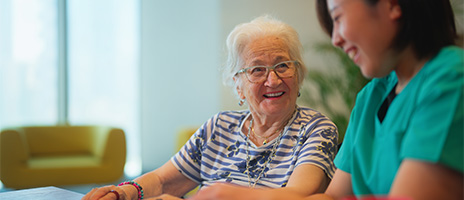 An elderly woman with white hair and glasses smiles while sitting beside a young caregiver in green scrubs. They appear to be having a friendly conversation at a table in a bright, modern room.