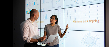 Two people stand in front of a large screen displaying charts, graphs, and the words Phenomic data mapping. One holds a laptop while they discuss the data, gesturing toward the display.