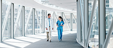 Two healthcare professionals, one in a white coat and one in blue scrubs, walk and talk in a bright, modern glass hallway lined with windows and steel beams.