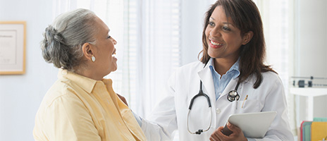 A doctor wearing a white coat and stethoscope smiles while talking to an older woman in a yellow shirt, offering reassurance. The interaction takes place in a bright, well-lit medical office.