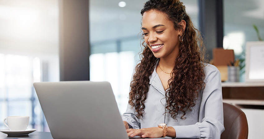 A woman with long curly hair, wearing a light blouse, sits at a desk smiling while working on her laptop in a modern, bright office environment, efficiently managing claims with denial management software for healthcare.