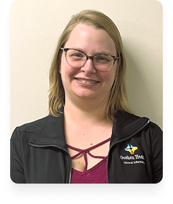 A woman with shoulder-length blonde hair and glasses smiles at the camera. She wears a black jacket with a Goshen Health Clinical Informatics logo and a maroon top underneath, standing in front of a light-colored wall.