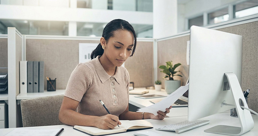 A woman sits at an office desk, writing in a notebook while holding papers related to HEDIS reporting, with a computer monitor, plant, and office supplies around her.